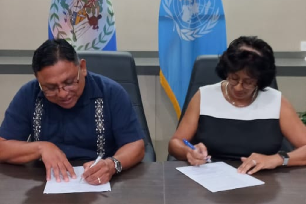 Two people seated at a desk sign an agreement in front of a United nations flag and the national flag of bELIZE