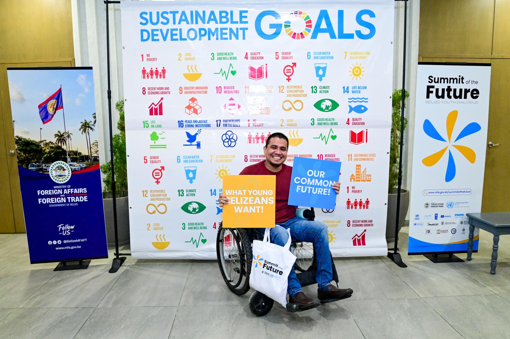 Man sitting in a wheelchair in front of SDG-themed screen that is flanked by Govt of Belize and Summit of the Future banners.