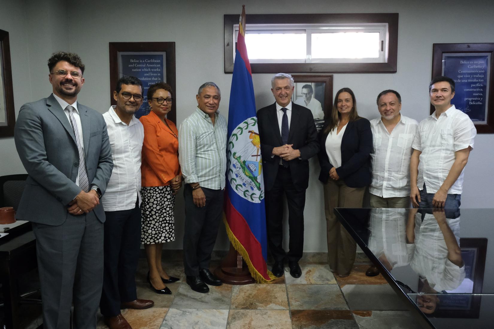 Eight persons standing in a semi-circle facing the camera.  A flag of Belize is between the persons standing at the top of the semi-circle