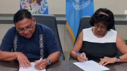Two people seated at a desk sign an agreement in front of a United nations flag and the national flag of bELIZE